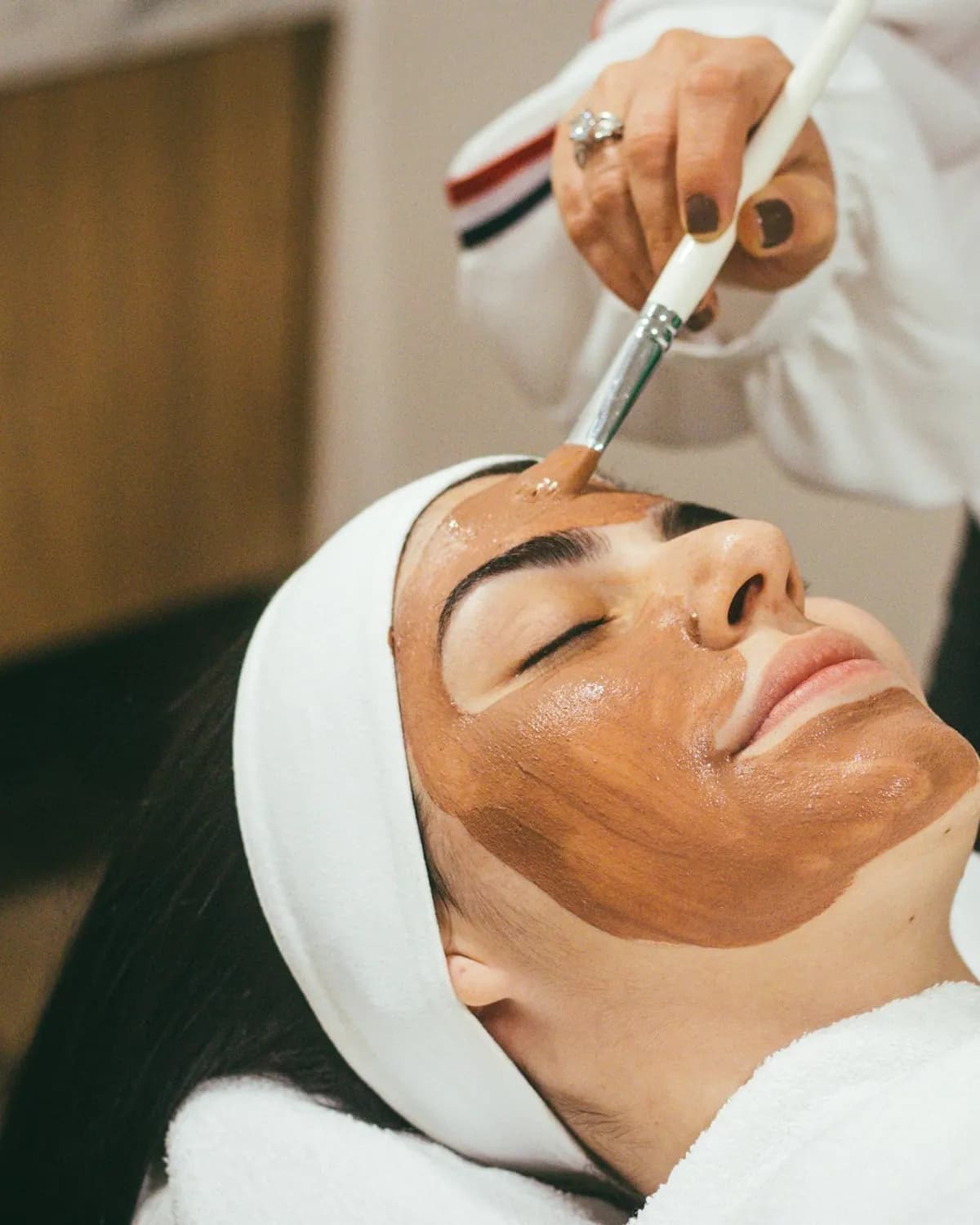 Mujer con los ojos cerrados y vincha blanca mientras le aplican una máscara facial con pincel