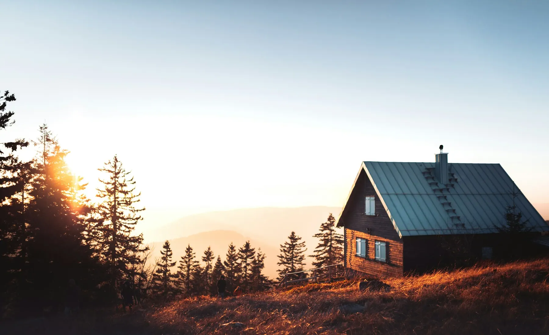 Cabaña de madera en una loma con pastizales y pinos, con las sierras en la bruma del atardecer