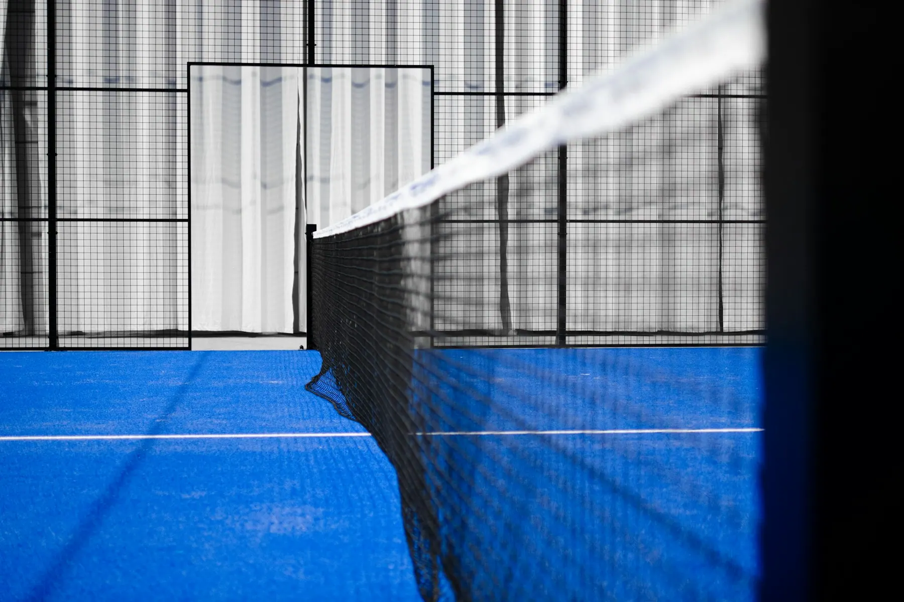 Cancha de pádel techada de césped azul, con la red en primer plano y el alambrado de fondo