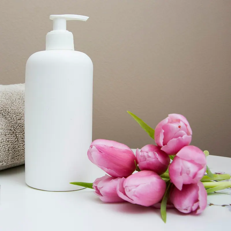 Frasco dosificador blanco de crema junto a una toalla enrollada, una vela y tulipanes rosas