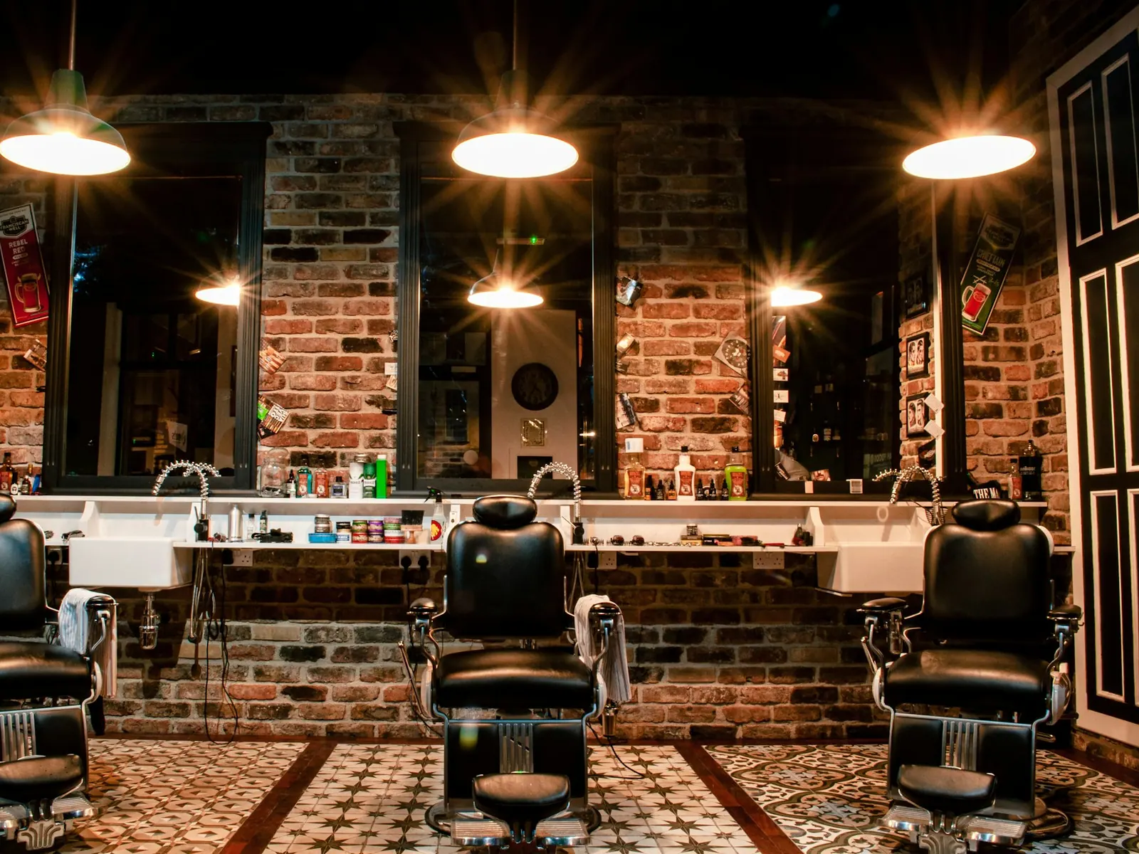Salón de la barbería: sillones de barbero de cuero negro frente a espejos, pared de ladrillo, lámparas colgantes y piso de mosaico